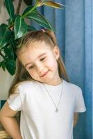 Smiling Little Girl in White Clothes with a Necklace Posing Near Houseplant, Looking at the Camera. photo