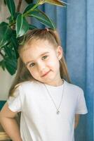 Smiling Girl in White Clothes with a Necklace Posing Near Houseplant, Looking at the Camera. photo
