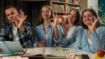 Four teenagers classmates reading book together preparing for exams showing ok gesture looking at camera smiling doing homework talking discuss good alright education studying pupils in school library photo