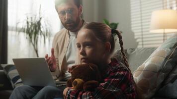 Caucasian family at home remote working dad using computer on sofa speaking to sad little girl child kid cuddling Teddy Bear conflict emotional stress childhood problem loneliness attention deficit photo