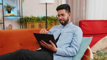 Happy Indian man freelancer work with documents on home couch reading information business papers prepare financial report. Young Hispanic guy accountant analyzing graph documents, calculate taxes. video