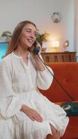 Smiling young woman making wired telephone call conversation with friends sitting on couch at home. Happy excited girl enjoying old-fashioned retro phone from 90s talking in living room. Vertical video