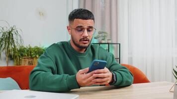 Sad displeased Arabian man use smartphone typing browsing, loses becoming surprised sudden lottery game results, bad news, fortune loss fail, deadline, virus. Young Indian guy at home office at table video