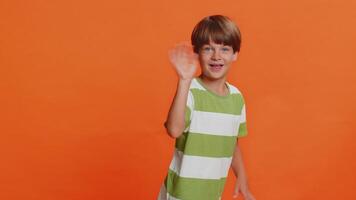 Young cute school boy 6 years old smiling friendly at camera and waving hands gesturing hello or goodbye, welcoming with hospitable expression. Preteen male child kid isolated on orange background video
