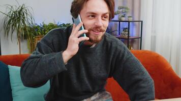 Phone call, good news, gossip. Happy surprised bearded man in pleasant conversation on smartphone, enjoying talking with friend indoors. Caucasian young guy sitting at table workplace home office room video