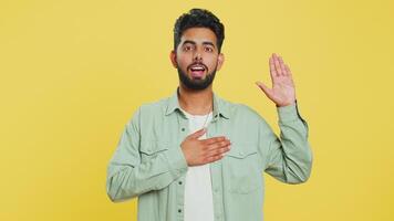 I swear to be honest. Sincere responsible Indian man raising hand to take oath, promising to be honest, to tell truth, keeping hand on chest, justice. Arabian guy isolated on yellow studio background video