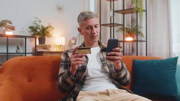 Caucasian man using credit bank card and smartphone while transferring money, purchases online shopping cashless order food delivery at home apartment indoors. Happy young guy sits in room on couch. video