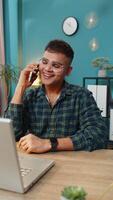 Indian man making phone call conversation with client or colleague friend. Arabian guy freelancer enjoying mobile loudspeaker talking gossip, rumors, good news at home office table workplace. Vertical video