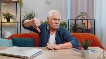 Dislike. Upset unhappy senior man showing thumbs down sign gesture, expressing discontent, disapproval dissatisfied bad work at home apartment indoors. Displeased elderly grandfather sits at table video