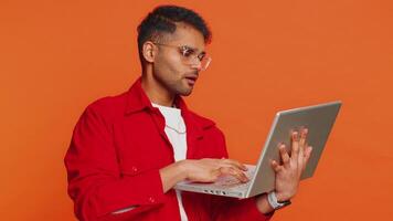 Displeased Indian man use laptop notebook typing browsing working, loses becoming surprised by lottery bad results fortune lost game fail computer virus, unlucky news. Arabian guy on orange background video