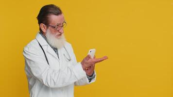 Senior doctor cardiologist man using smartphone showing pointing empty place, advertising application area, positive feedback, copy-space. Apothecary pharmacy grandfather isolated on yellow background video