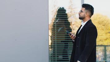 Side view of Indian businessman using smartphone typing e-mail answering messages chatting online looking mobile screen app. Arabian Hindu freelancer guy walking downtown city street. Business people video