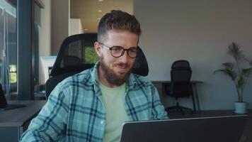Young, bearded office worker wearing glasses sits in his chair after hours, savoring glass of whiskey while enjoying moment of solitude and relaxation in quiet evening. Speech of alcohol at workplace video