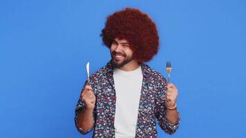 Ready to eat. Hungry man with Afro hairstyle wig waiting for serving dinner dishes with with restlessness holding cutlery fork knife, will appreciate delicious restaurant meal. Guy on blue background video