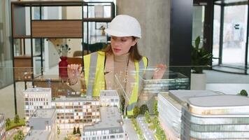 Confident young female architect in hardhat and reflective vest analyzing looking at business office complex architectural model. Woman in protective clothing designer of buildings working in office. video