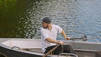 Young man is fisherman, trying to carefully row his rowboat to the shore on a pond in the summer. He uses oars to move the boat and moor it safely. video