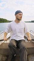 Attractive man, fisherman with beard and dockworker's hat, is rowing boat with two hands in middle of the river during the summer. video