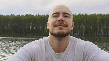 Friendly young man with beard and bald head, who looks strong and brave, is rowing rowboat with oars on river in summer, against backdrop of forest on shore. Portrait. video