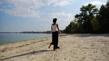 Happy adult woman jogging on sandy beach near shoreline at sunny day. Beautiful lady running on seashore with calm waves at background. Concept of summer vacation or holiday. Slow motion video