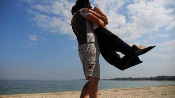 Carefree lovers having fun together on seashore at summer vacation. Man taking his beloved in air, turning around and kissing her at coastline. Happy couple spending time at sandy beach. Slow mo video