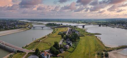 Aerial view of Nijmegen, Netherlands, showcasing the Waal River, De Oversteek, Waalbrug, green fields, residential areas, and scattered clouds in the sky. photo