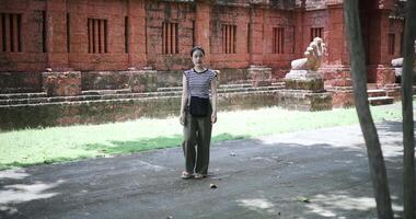 Handheld shot, Relaxed woman wearing a striped shirt and green pants walking around, admiring the natural scenery and ancient brick structures, Enjoy holiday travel. video