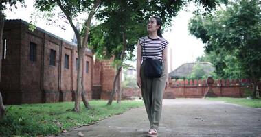 Handheld low angle view shot, Young Asian woman in a striped shirt and green pants walks along a shady tree-lined path. Enjoy holiday travel. video