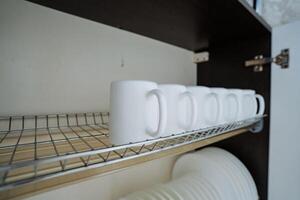 A neat row of pristine white mugs, carefully arranged, are sitting on a stylish wire rack in the kitchen cabinet, easily accessible for everyday use or special occasions photo
