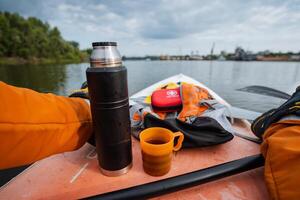 A person is sitting comfortably on a kayak that is floating on the water, holding a thermos in one hand and a steaming cup of coffee in the other, enjoying the serene surroundings photo