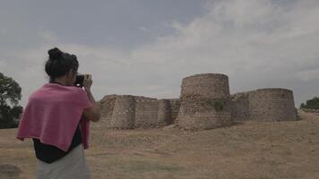 Tourist capturing photo of historic stone fortress walls on dry grassy field under cloudy sky video