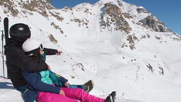 Relaxed couple in ski gear sitting together on snowy mountain slope. Pointing at distant peaks, enjoying breathtaking alpine view, symbolizing adventure, freedom, romance, travel inspiration. video