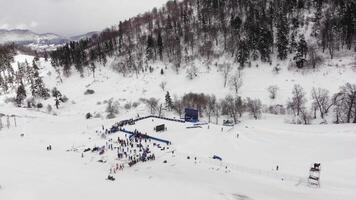 Aerial drone view of a snowy mountain sports competition with crowds gathered at the base and athletes on slopes during winter. video