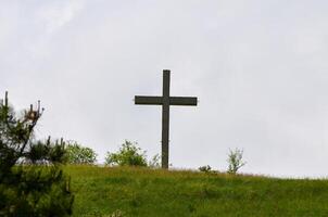 A wooden cross is standing on a hillside in a grassy field. The sky is cloudy, and the cross is the only visible object in the scene photo