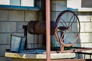 A rusty old water pump is hanging from a chain. The chain is attached to a bucket. The bucket is empty photo