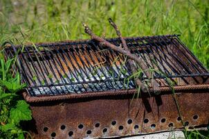 A rusty grill with a few sticks on it. The grill is on a grassy area. The sticks are on the grill and are not burning photo