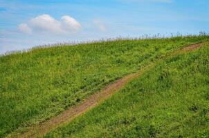 A grassy hill with a dirt path leading down it. The sky is clear and blue photo