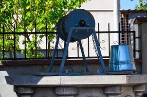 A rusty metal object is sitting on a ledge next to a bucket. The object appears to be a pulley or a crank, and the bucket is made of metal. The scene has a somewhat industrial or mechanical feel photo