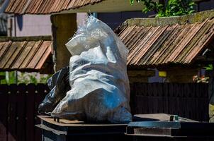 A white bag is on top of a trash can. The bag is full of trash and is covered in plastic photo