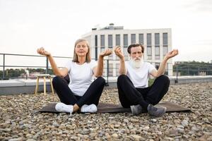 Senior man and woman practicing meditation together, sitting in lotus pose on rooftop stones. Emitting calmness, peaceful energy. Comfortable clothing fits outdoor artistic environment urban buildings photo