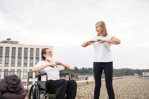 Elderly woman assisting man in wheelchair practicing exercises on rooftop, promoting fitness and well-being. Man and woman demonstrating teamwork, inclusivity, and active lifestyle. photo