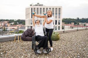 Senior woman assists elderly husband exercising with dumbbells while wheelchair bound, outdoor setting showcases mutual support and healthy lifestyle promotion despite physical limitations. photo
