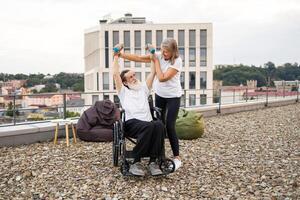 Senior woman assisting wheelchair-bound husband with rehabilitation exercises using dumbbells on rooftop. Bright day enhancing supportive, emotional interaction, emphasizing recovery, togetherness. photo