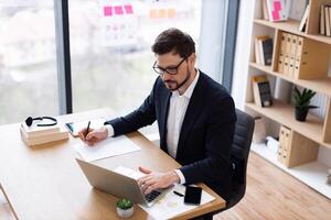 Young adult male in formal attire working in office, using laptop, taking notes on paper, conducting business task. Office environment has natural light, bookshelves, and work materials. photo