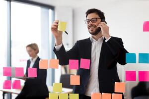 Businessman wearing glasses writing on sticky notes on transparent board while on phone. Professional setting suggesting planning organization and productivity. Female colleague in background working. photo