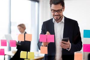 Adult male organizing tasks with colorful sticky notes on glass board, holding smartphone. Office backdrop with modern workplace vibes. photo