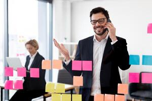 Adult man in formal attire speaking over smartphone near glass board with colorful post-it notes. Office setting with second person in background working on laptop. Collaboration and planning concept. photo