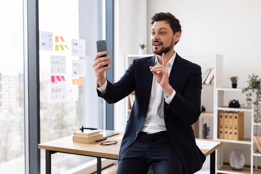 Man in business attire engaging in communication using smartphone in office environment. Professional workspace includes desk, documents, and shelves. Represents modern technology and virtual meetings photo