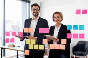 Two adult business professionals using sticky notes on glass board for brainstorming session. Concept of teamwork collaboration and strategizing in office setting featuring multicultural human models photo