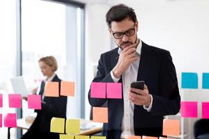 Confident adult man in formal suit analyzing colorful sticky notes on glass in modern workspace. Assisting female colleague visible in background. photo