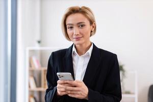 Elegant adult woman wearing formal suit standing indoors holding smartphone looking confident. Represents business communication, technology usage. Professional setting, light and welcoming workspace photo